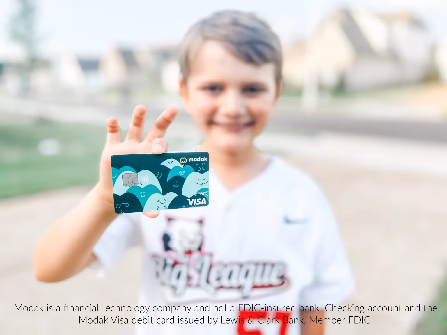 Smiling boy in a baseball jersey holding a teal Modak debit card outdoors