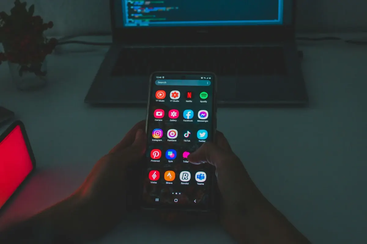 Hand holding a phone with app icons in front of a laptop