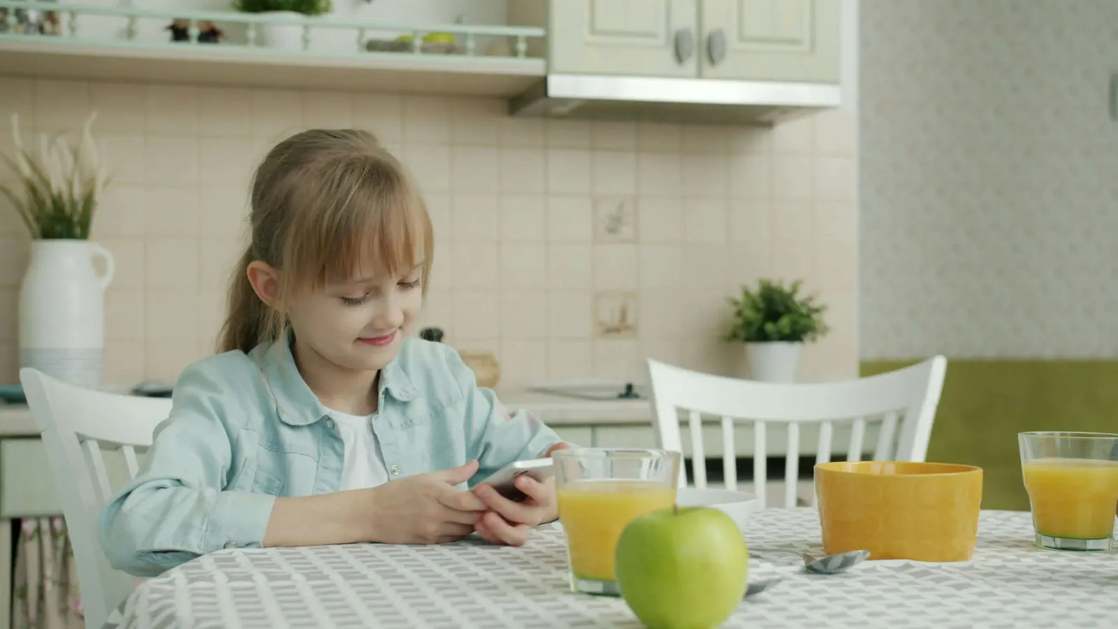 Girl at a kitchen table looking at her phone next to juice and an apple