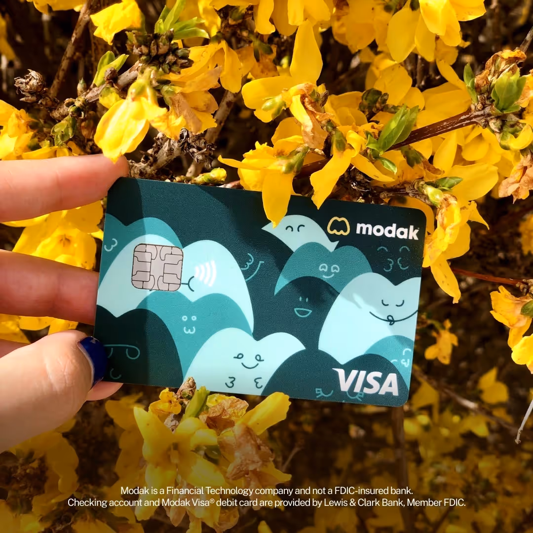 Teal Modak Visa debit card held up against yellow flowers