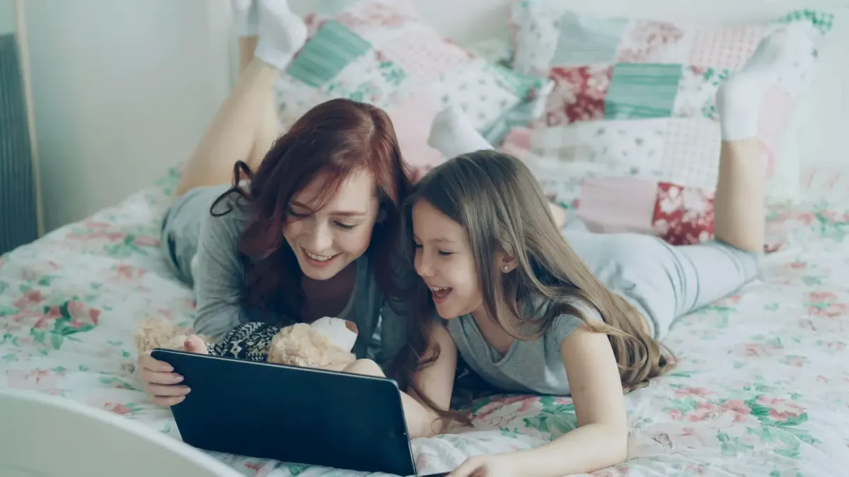 Mother and daughter smiling while looking at a tablet together on a bed