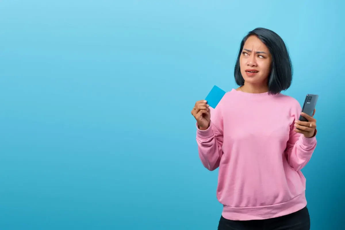 Woman in pink sweatshirt holding a blue card and phone with a worried look